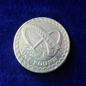 Puede incluir: Una moneda británica de una libra de plata con un diseño del Millennium Dome, una gran estructura blanca en forma de cúpula con un anillo alrededor de la base. La moneda tiene las palabras "ONE POUND" inscritas alrededor del borde.