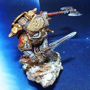 May include: A detailed miniature figure of a warrior in ornate armor, kneeling on a snowy base. The figure wields a large double-bladed axe and a long sword. The armor is gold, silver, and red, with wolf-themed shoulder pads.
