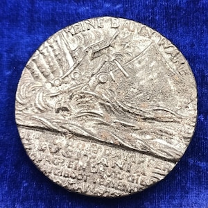 Puede incluir: Una medalla redonda de color plateado con imágenes en relieve de un barco en el mar y texto. La superficie de la medalla tiene un aspecto texturizado. El texto de la medalla está en alemán.