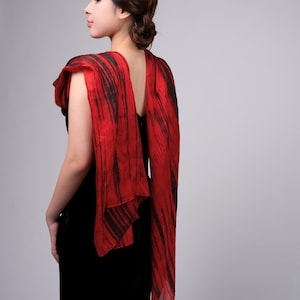 May include: A red and black patterned scarf draped over a black dress. The scarf features a vibrant red base with black vertical streaks, creating a striking contrast. The fabric appears lightweight and flowing, adding elegance.