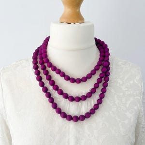 Può includere: Una collana a tre fili con grandi perle rotonde, viola scuro. La collana è esposta su un manichino bianco con collo in legno. Il manichino indossa una camicetta bianca strutturata.