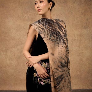 Puede incluir: Un vestido negro con bordado floral y un chal teñido anudado. El chal presenta un patrón de líneas negras sobre un fondo beige. El vestido tiene una silueta ajustada.