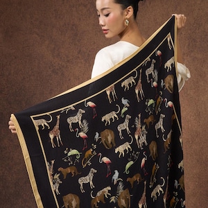 May include: Black square scarf with a gold border featuring a safari animal print. The scarf displays illustrations of giraffes, zebras, leopards, elephants, and flamingos. The scarf is held up by a person.