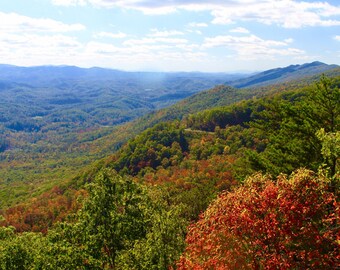 May include: A panoramic view of a mountain range with fall foliage. The trees in the foreground are a mix of green, yellow, and red. The mountains in the distance are blue and hazy. The sky is blue with white clouds.