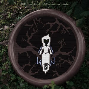 May include: A cross-stitch pattern featuring a white hooded figure with yellow eyes perched on a brown tree branch against a black background. The design is circular and includes the text PDF download . 100% human made at the top of the frame.