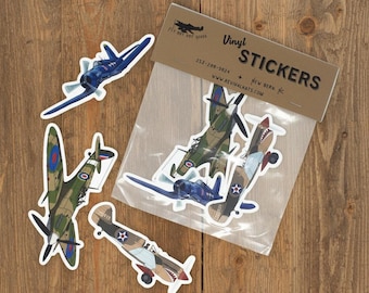 May include: A collection of vinyl stickers featuring illustrations of vintage military aircraft. The stickers are packaged in a clear plastic sleeve with a brown cardboard header labeled Vinyl STICKERS, Fly Boy Dry Goods, and contact details.