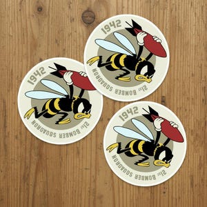 May include: Three circular stickers featuring a cartoon bee with yellow and black stripes, white wings, and white gloves, carrying a red bomb. The text 1942 and 21st BOMBER SQUADRON is printed in a circular arc around the central bee illustration.
