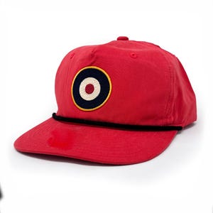 May include: A bright red baseball cap featuring a circular embroidered patch on the front. The patch displays a navy blue outer ring, a white middle ring, and a red center, all outlined in yellow. A thin black cord runs across the brim base.
