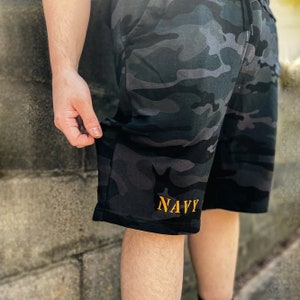 May include: Black and gray camouflage print shorts with a gold "NAVY" embroidered on the left leg.