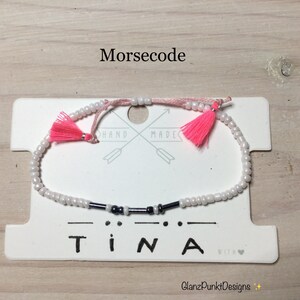 May include: White beaded bracelet with black beads spelling out "TINA" in Morse code. The bracelet has pink tassels on each end and a white card with the words "HAND MADE" and "with love" written on it.