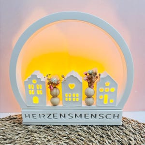 Könnte beinhalten: Ein weißes dekoratives Licht mit einem kreisförmigen Rahmen und einem warmen gelben Schein. Es hat vier beleuchtete hausförmige Strukturen mit kleinen Vasen, die getrocknete Blumen halten. Das Wort "HERZENSMENSCH" wird am Sockel angezeigt.