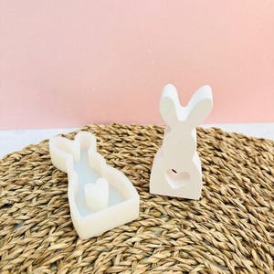 Könnte beinhalten: Zwei weiße, hasenförmige Objekte. Eines ist eine Form mit einer herzförmigen Kerze im Inneren, und das andere ist ein fester Hase mit einem herzförmigen Ausschnitt. Die Objekte befinden sich auf einer gewebten Matte mit rosa Hintergrund.