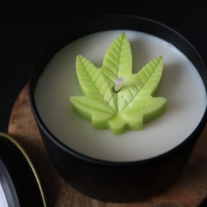 Könnte beinhalten: Eine weiße Kerze in einer schwarzen Dose mit einem grünen Wachsdesign in Form eines Cannabisblattes. Die Kerze steht auf einer Holzoberfläche. Der Deckel der Dose ist unten links sichtbar.