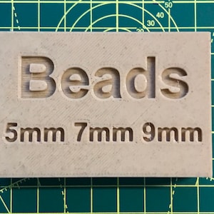 May include: A beige rectangular label with the word "Beads" in large, bold, sans-serif font. Below the word "Beads" are the sizes "5mm 7mm 9mm" in a smaller, sans-serif font.