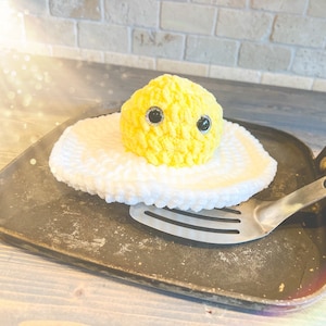 May include: A yellow crocheted egg with black eyes sits on a white crocheted fried egg in a black skillet with a silver spatula.
