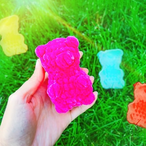 May include: Four gummy bear shaped soaps in different colours: yellow, pink, blue and orange. The pink gummy bear soap is being held in a hand.
