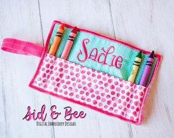 May include: A pink and turquoise fabric crayon holder with a polka dot pattern and the name "Sadie" embroidered on the front. The holder has a pink strap and is filled with crayons.