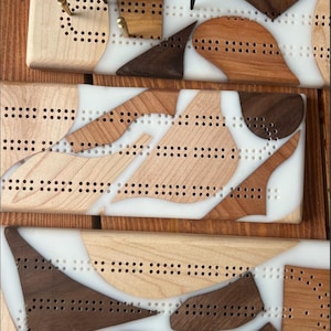 May include: Three wooden cribbage boards with white epoxy resin inlays. The boards are made of maple, walnut, and cherry wood. Each board has a unique design with different shapes and sizes of wood inlays.