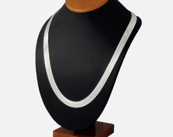 Puede incluir: Un collar de plata con diseño de espiga exhibido en un busto de joyería negro con base y cuello de madera. El collar tiene un diseño plano y tejido, que refleja la luz. El busto está sobre un fondo neutro.