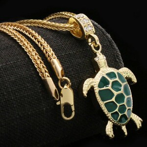 Könnte beinhalten: Goldfarbene Halskette mit einem Schildkrötenanhänger. Der Anhänger hat einen grünen Emaille-Schild und goldfarbene Details mit kleinen, klaren Steinen. Die Kette hat ein geflochtenes Design und einen Karabinerverschluss.