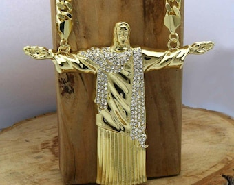 May include: Gold-toned pendant necklace featuring a detailed depiction of Christ the Redeemer with outstretched arms. The pendant is adorned with sparkling crystals and hangs from a thick, gold chain. The necklace is displayed against a wooden background.