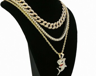 May include: A collection of gold-toned necklaces displayed on a black mannequin. The necklaces include a thick chain, a tennis chain, a rope chain, and a pendant featuring a shark design with red accents. The jewelry is adorned with clear crystals.