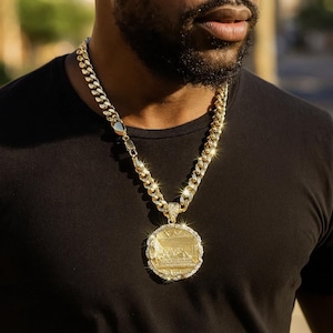 Può includere: Una collana a catena color oro con un grande pendente rotondo. Il pendente presenta un design dettagliato con un'immagine centrale ed è incrostato di piccole pietre scintillanti. La catena ha un design a maglie spesse.