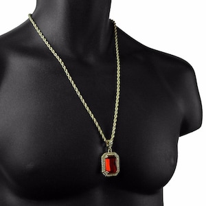 May include: Gold-coloured rope chain necklace featuring a rectangular red gemstone pendant. The pendant is framed with small, clear stones. The necklace is displayed on a black mannequin.