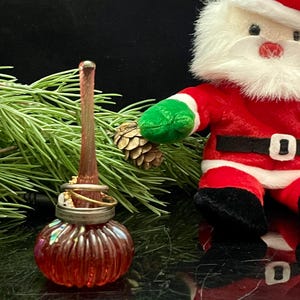 May include: A red glass perfume bottle with a ribbed design, topped with rings and a long, slender applicator. A small Santa Claus figure and pine needles are in the background, suggesting a holiday theme.