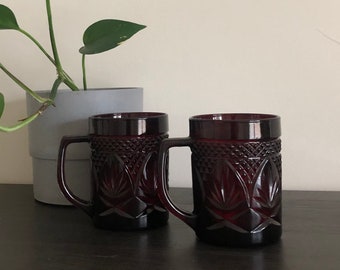 May include: Two red glass mugs with a textured design. The mugs have handles and are sitting on a dark brown surface.