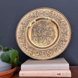May include: A vintage brass decorative plate with an embossed scene of figures in period clothing. The plate has an ornate border with repeating figures and floral designs. The plate is displayed on a stack of books, with a potted plant in the foreground.