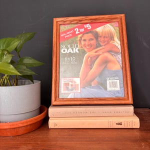 May include: A solid oak 8x10 photo frame with a picture of a woman and child. The frame has a warm brown finish and is displayed on top of two books. The frame has a sticker that says "2 for $5".
