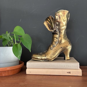 May include: A vintage brass boot sculpture with a high heel and button details. The boot is displayed on top of two books with neutral-colored covers. A potted plant with green leaves is to the left of the boot.