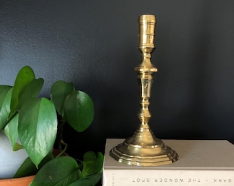 May include: A gold metal candlestick with a simple design. The candlestick is sitting on top of two books with the text "BANK. THE WONDER SPOT" visible.