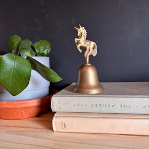 May include: A brass hand bell featuring a unicorn figurine on the handle. The bell has a smooth, conical base and a detailed unicorn topper. It is positioned on top of two stacked books, one of which is titled The Wonder Spot by Viking.