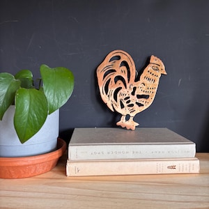 May include: A copper-colored metal wall hanging shaped like a rooster with intricate cut-out patterns throughout its body. The rooster features a textured surface and is positioned upright behind two stacked hardcover books on a wooden surface.