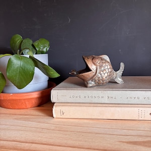 May include: A metallic, copper-toned fish figurine with a wide-open mouth and textured scale detailing, positioned on top of two stacked hardcover books. The fish has a prominent tail fin and a rounded body shape, resting on a flat surface.