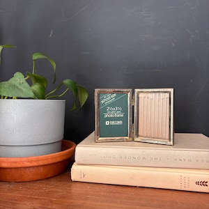 May include: A hinged, school-size photo frame with a gold-toned border, open to display a green backing and a blank space. The frame is labeled "2 1/2 x 3 1/2" and "non-glare glass." It rests on a stack of books.