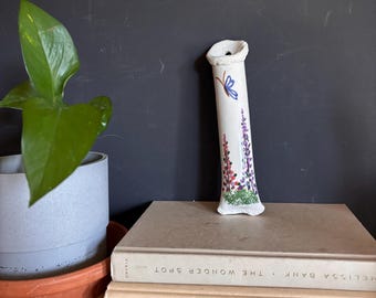 May include: A tall, slender white ceramic bud vase with a textured rim. The vase features a hand-painted design of a blue butterfly, purple and pink lupine flowers, and green foliage at the base. It stands upright on a stack of two hardcover books.