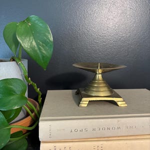 May include: A brass candle holder with a square base and a conical stem, topped with a shallow dish. The candle holder sits on a stack of books with the titles "The Wonder Spot" and "At the Entrance to the Garden of Eden". A green plant is in the background.