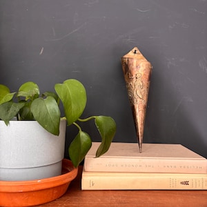 May include: A copper-colored, cone-shaped wall vase with an embossed design of a figure. The vase is displayed on a stack of books next to a potted plant with green leaves. The vase has a small hole at the top for hanging.
