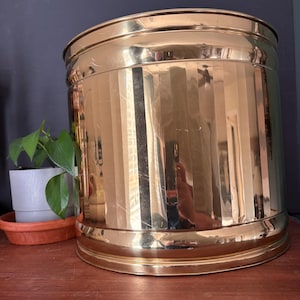 May include: A large, cylindrical, polished brass planter featuring vertical fluted detailing along the body. The metallic surface is highly reflective, showing light and surrounding objects. It has a smooth, rounded rim and a wide, flat base.