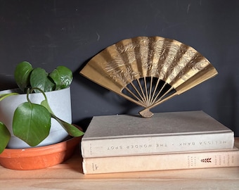 May include: A decorative brass hand fan featuring an embossed floral and bird design, displayed standing upright on top of two stacked hardcover books. The fan has a metallic gold finish and a semi-circular shape with visible radiating ribs.