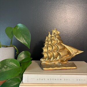 May include: A gold metal ship bookend with intricate details. The bookend is sitting on top of two books with the titles "Bank The Wonder Spot" and "At the Entrance to the Garden of Eden" visible.