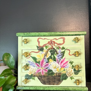 Może przedstawiać: Jasnozielone pudełko na biżuterię z czterema szufladami, ozdobione malowanym wzorem kosza kwiatowego. Kosz zawiera różowe lilie, białe róże i inne kwiaty. Złote uchwyty szuflad w kształcie pszczół dodają dekoracyjnego akcentu.