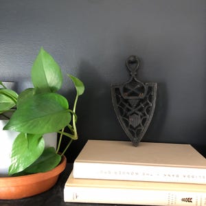 May include: A black cast iron trivet with a decorative design. The trivet is shaped like a shield and has a handle on the top. The trivet is sitting on a stack of books.