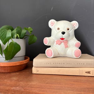 Puede incluir: Una figura de oso de peluche de cerámica blanca con detalles en rosa, incluyendo un lazo, patas y detalles en las orejas. El oso está colocado sobre dos libros en una superficie de madera, con una planta en maceta en el fondo.