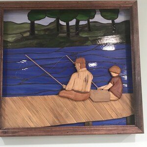 May include: A framed wood carving of two figures fishing from a wooden dock. The figures are a man and a boy, both wearing brown clothing. The background is a stained glass depiction of a lake with blue water and green trees.