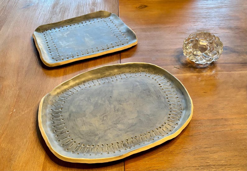 Op de afbeelding: Twee keramische serveerschalen met een grijze binnenkant en een gele rand. De schalen hebben een decoratief stippenpatroon rond de rand. Er is ook een kleine, heldere glazen votiefhouder aanwezig.