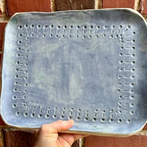 Op de afbeelding: Een rechthoekige keramische serveerschaal met een lichtblauwe glazuur. De schaal heeft een decoratieve rand van kleine, gelijkmatig verdeelde gaatjes. De schaal wordt door een hand tegen een bakstenen muur gehouden.
